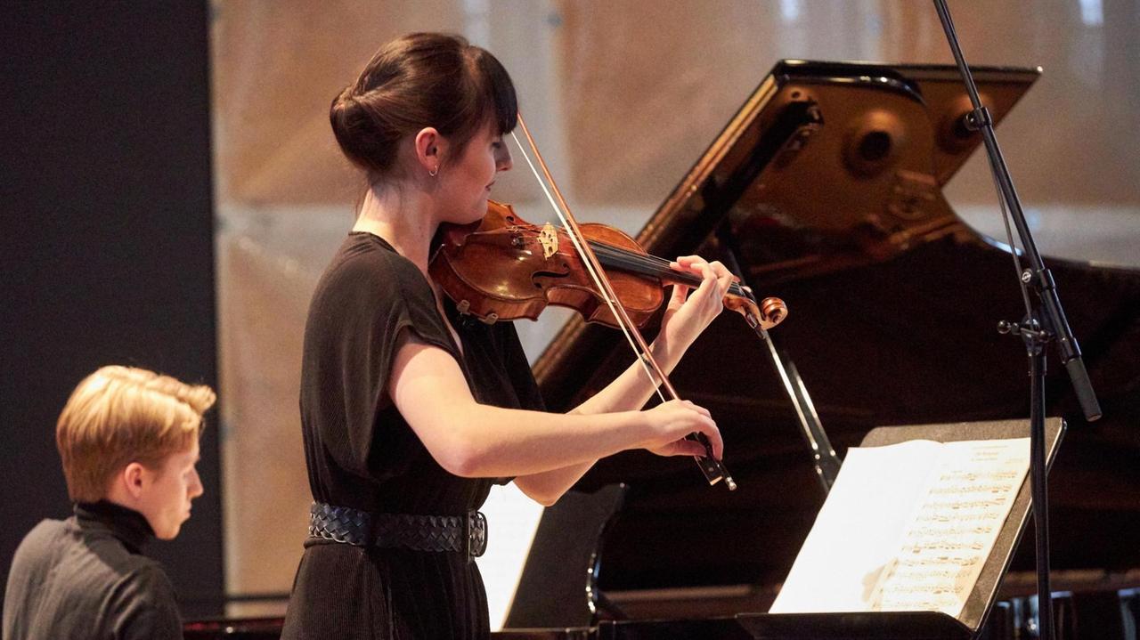 Von der Seite ist die Geigerin spielend vor ihrem Notenpult zu sehen, im Hintergrund sitzt der Pianist am offenen Flügel. Von der Seite ist die Geigerin spielend vor ihrem Notenpult zu sehen, im Hintergrund sitzt der Pianist am offenen Flügel.
