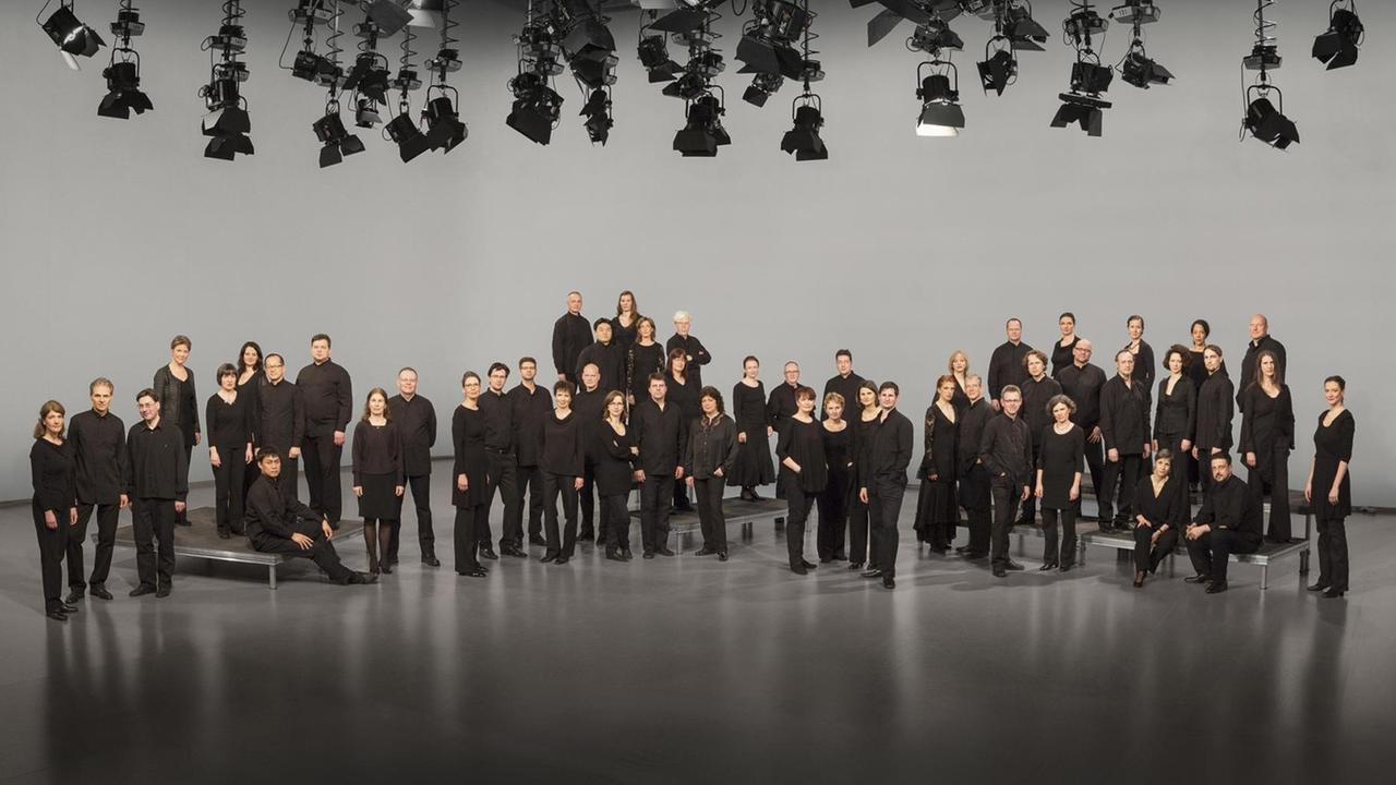 Der schwarz gekleidete Chor steht auf einer Bühne, von deren Himmel zahlreiche Scheinwerfer ins Bild ragen. Der schwarz gekleidete Chor steht auf einer Bühne, von deren Himmel zahlreiche Scheinwerfer ins Bild ragen.