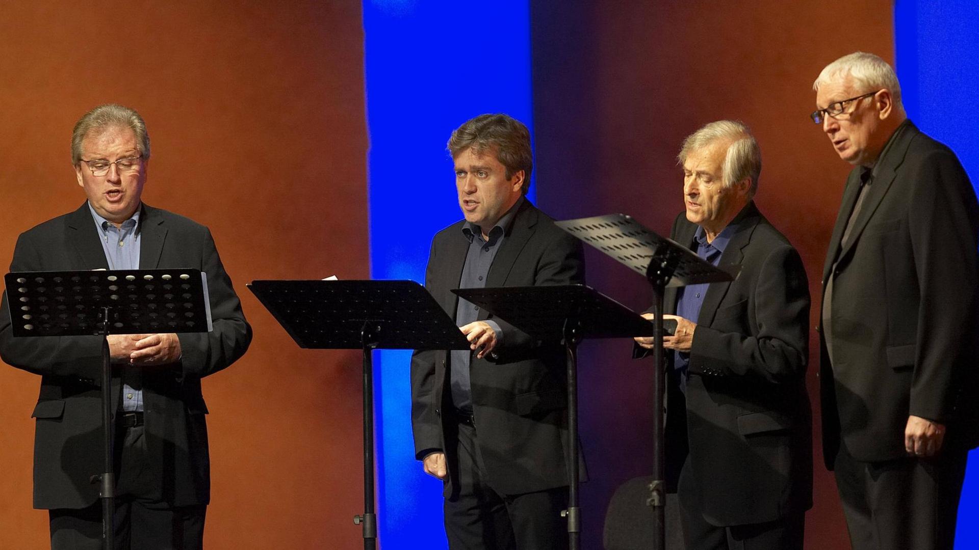 Vier ältere Herren stehen hinter Notenpulten und singen auf einer Bühne. Vier ältere Herren stehen hinter Notenpulten und singen auf einer Bühne.