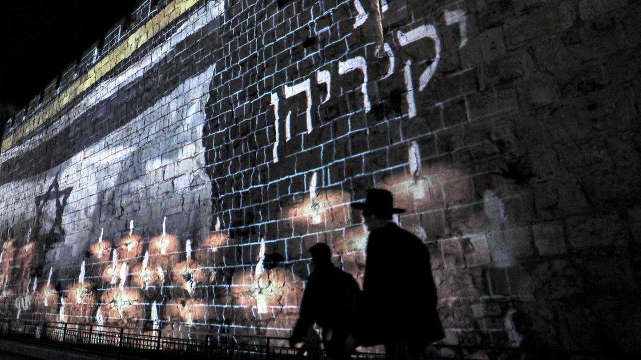 Eine israelische Flagge, die vor brennenden Kerzen weht, ist an die alte Stadtmauer von Jerusalem projiziert. Davor sind die Silhouetten von zweit orthodexen Männern zu sehen. Am 2. Mai hat Israel einen nationalen Trauertag für die Opfer einer Massenpanik während des Lag-BaOmer-Festes am Berg Meron ausruft. Das tödliche Gedränge am Berg Meron im Norden Israels ist eine der schlimmsten Katastrophen in Friedenszeiten seit der Gründung der Nation im Jahr 1948.