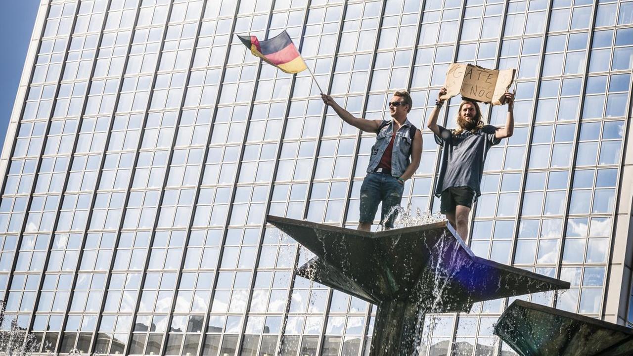 Zwei Demonstranten bei einer "Hygienedemo"protestieren auf dem Brunnen der Völkerfreundschaft auf dem Alexanderplatz in Berlin. Einer hält ein Schild: "Gate's noch?", ein anderer schwingt die Deutschlandfahne, 9. Mai 2020.