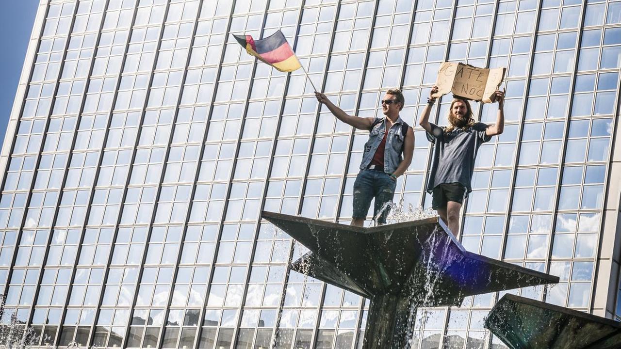 Zwei Demonstranten bei einer "Hygienedemo"protestieren auf dem Brunnen der Völkerfreundschaft auf dem Alexanderplatz in Berlin. Einer hält ein Schild: "Gate's noch?", ein anderer schwingt die Deutschlandfahne, 9. Mai 2020. Zwei Demonstranten bei einer "Hygienedemo"protestieren auf dem Brunnen der Völkerfreundschaft auf dem Alexanderplatz in Berlin. Einer hält ein Schild: "Gate's noch?", ein anderer schwingt die Deutschlandfahne, 9. Mai 2020.