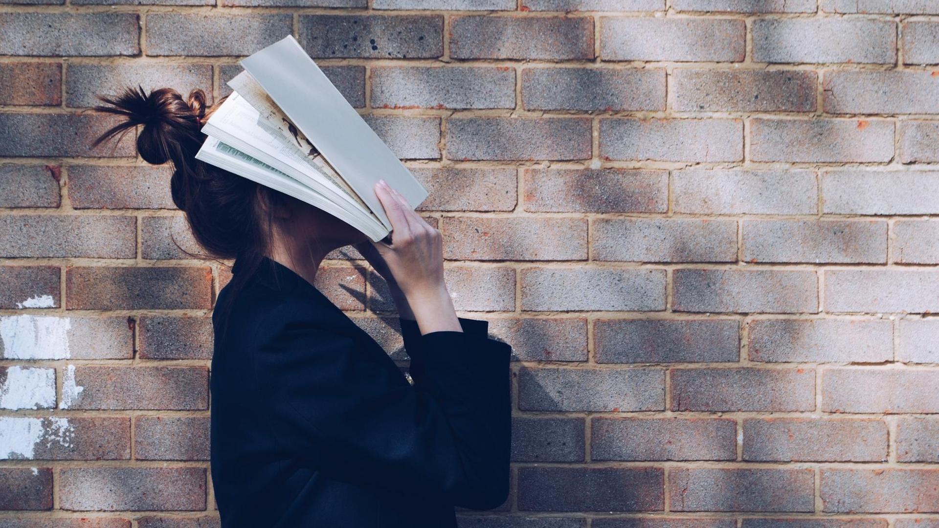 Eine Frau steht vor einer Mauer und drückt ihr Gesicht in ein offenes Buch. Die Szene hat etwas verzweifeltes.