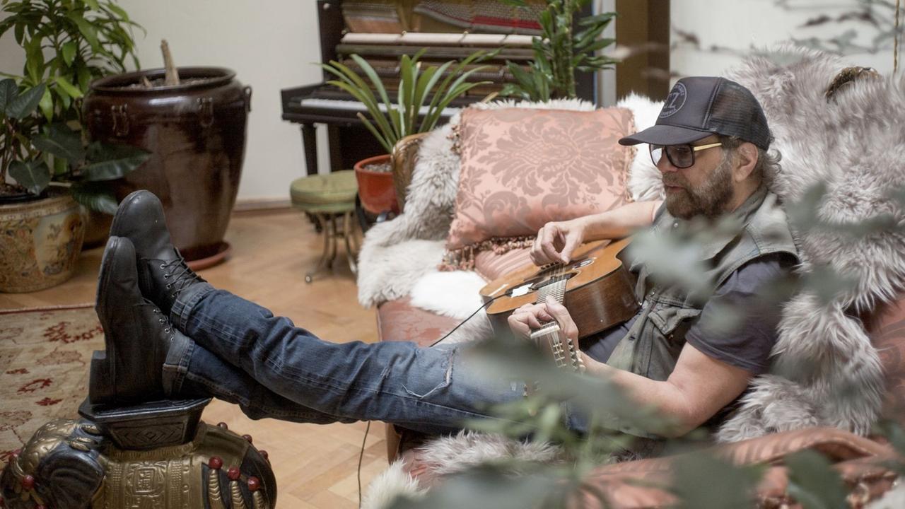 Ein Mann mit Gitarre sitzt auf einem Sofa und hat die Füsse auf einen Hocker gelegt. Im Hintergrund steht ein Klavier und mehrere Pflanzen.