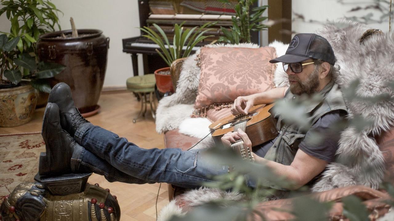 Ein Mann mit Gitarre sitzt auf einem Sofa und hat die Füsse auf einen Hocker gelegt. Im Hintergrund steht ein Klavier und mehrere Pflanzen. Ein Mann mit Gitarre sitzt auf einem Sofa und hat die Füsse auf einen Hocker gelegt. Im Hintergrund steht ein Klavier und mehrere Pflanzen.