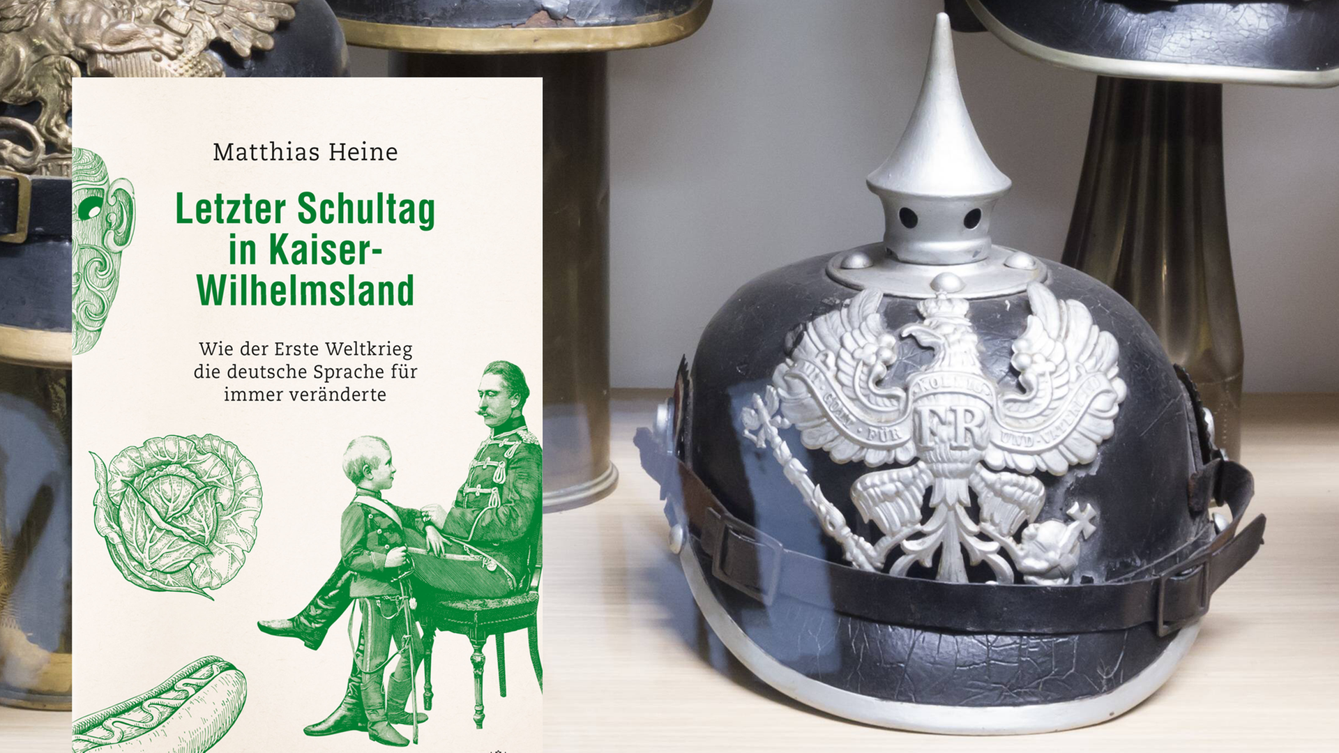 Eine Pickelhaube in einer Ausstellungsvitrine. Dafür das Cover zu Matthias Heines Buch "Letzer Schultag in Kaiser-Wilhelmsland" auf dem eine Zeichnung des Schulalltags im frühen 20. Jahrhundert zu sehen ist.