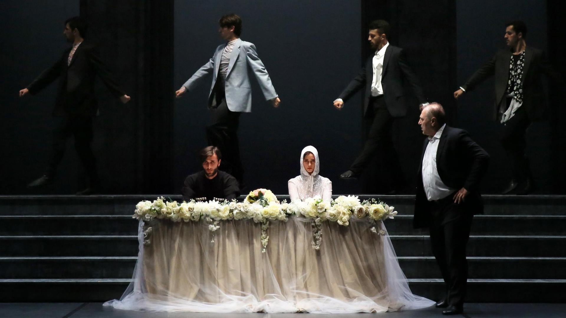 Zwei Mitglieder des Tanzensembles (LA)HORDE sitzen wie Braut und Bräutigam hinter einem Blumenpult, vier Männer in Anzügen tanzen hinter ihnen, ein Mann im Anzug steht rechts neben ihnen.