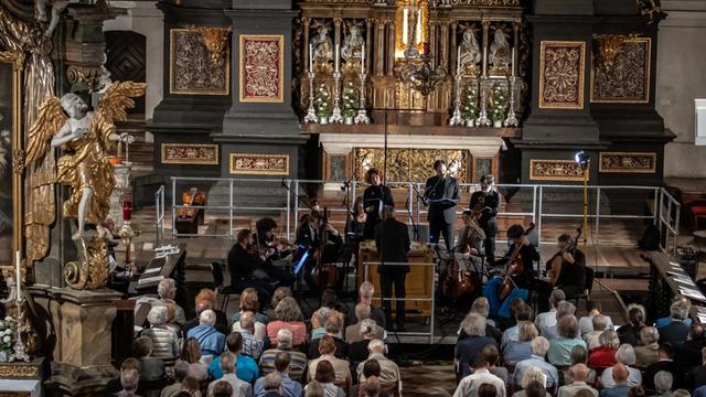 Ein Sängerensemble singt in einer barocken Kirche ein Live-Konzert. Man sieht die Hinterköpfe der Menschen im Publikum. Die Sänger sind alle schwarz gekleidet.