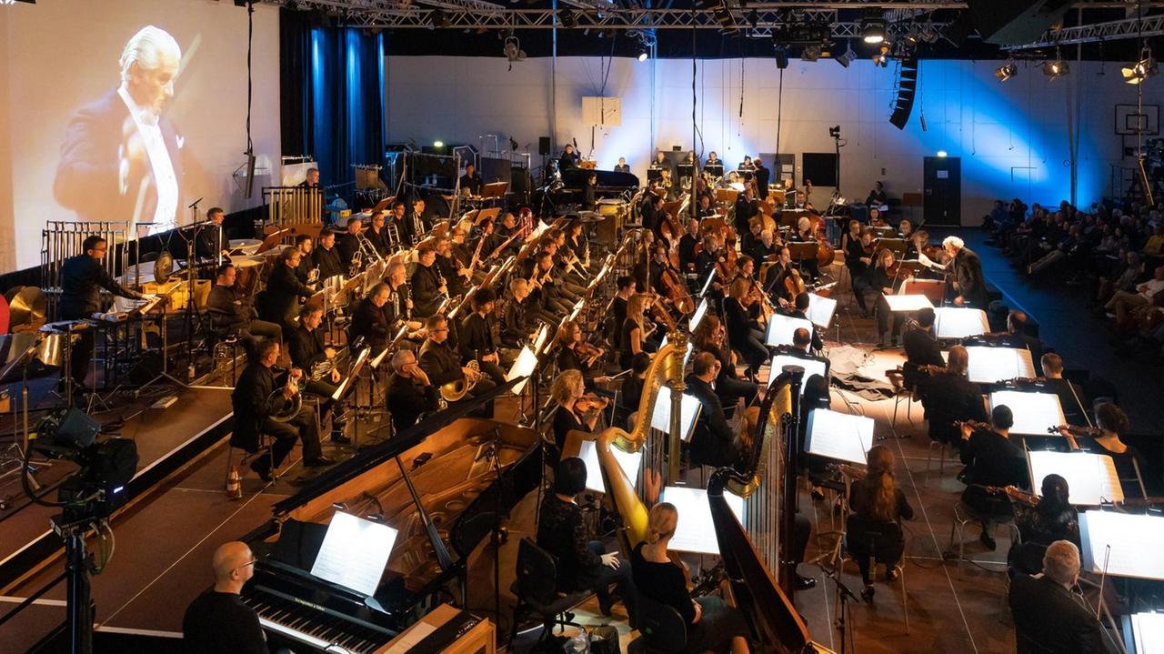 Ein großes Orchester sitzt auf einer Bühne, dahinter eine Leinwand, auf der eine Großaufnahme eines Dirigenten zu sehen ist.