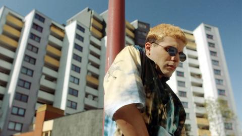 Filmstill aus "Futur Drei": Eine junger Mann mit blondierten Haaren und einer Sonnenbrille kramt in der Tasche seiner weißen Jeans. Hinter ihm ragt ein Hochhaus.