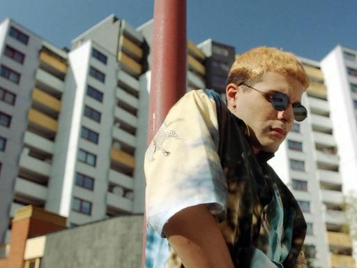 Filmstill aus "Futur Drei": Eine junger Mann mit blondierten Haaren und einer Sonnenbrille kramt in der Tasche seiner weißen Jeans. Hinter ihm ragt ein Hochhaus.