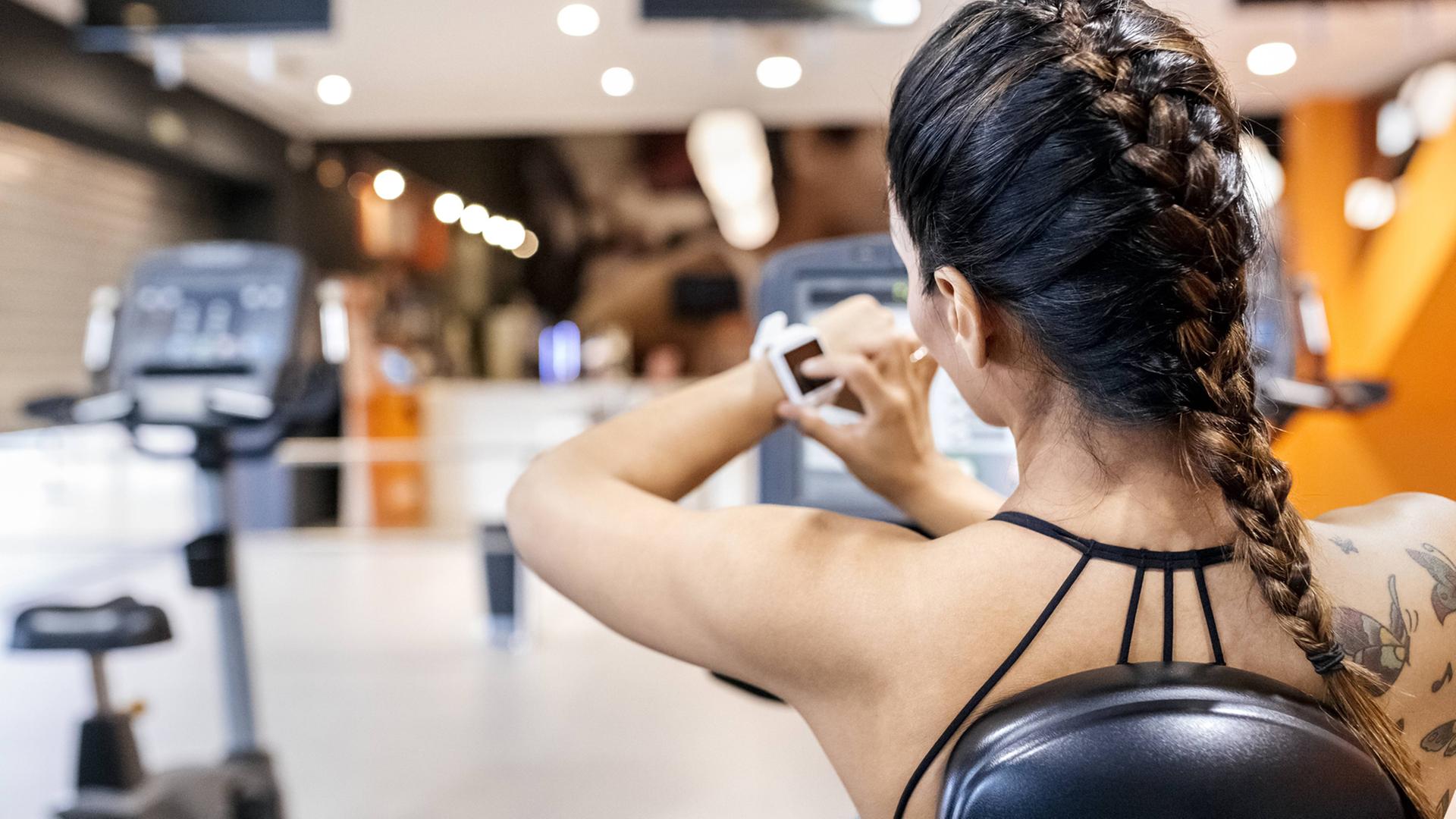 Rückansicht eine Frau im Fitnesstudio, die auf ihre Smartwatch schaut. Auf der Schulter rechts hat sie mehrere Tätowierungen. Rückansicht eine Frau im Fitnesstudio, die auf ihre Smartwatch schaut. Auf der Schulter rechts hat sie mehrere Tätowierungen.