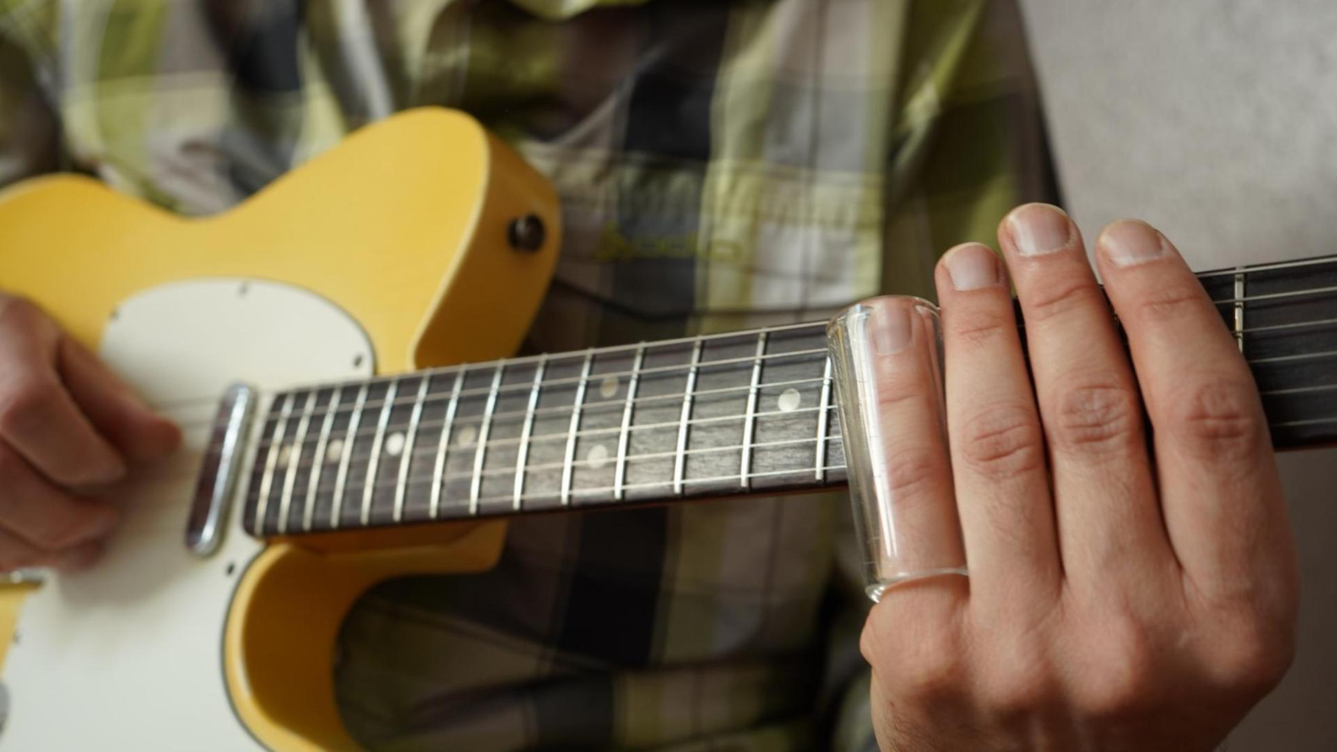 Ein Mann hat ein Glasrohr auf seinem Ringfinger stecken und greift mit der Hand den Hals einer gelben Gitarre, einer Fender Telecaster.