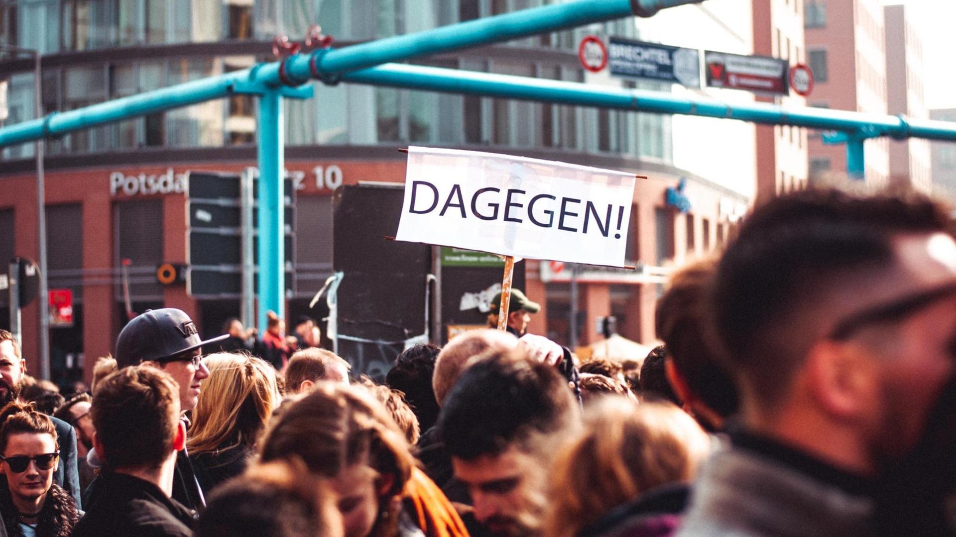 Köpfe einer Menschenmenge am Potsdamer Platz in Berlin. Es handelt sich um eine Demo. Zwischen den Köpfen ragt ein Schild mit der Aufschrift "Dagegen" hervor.