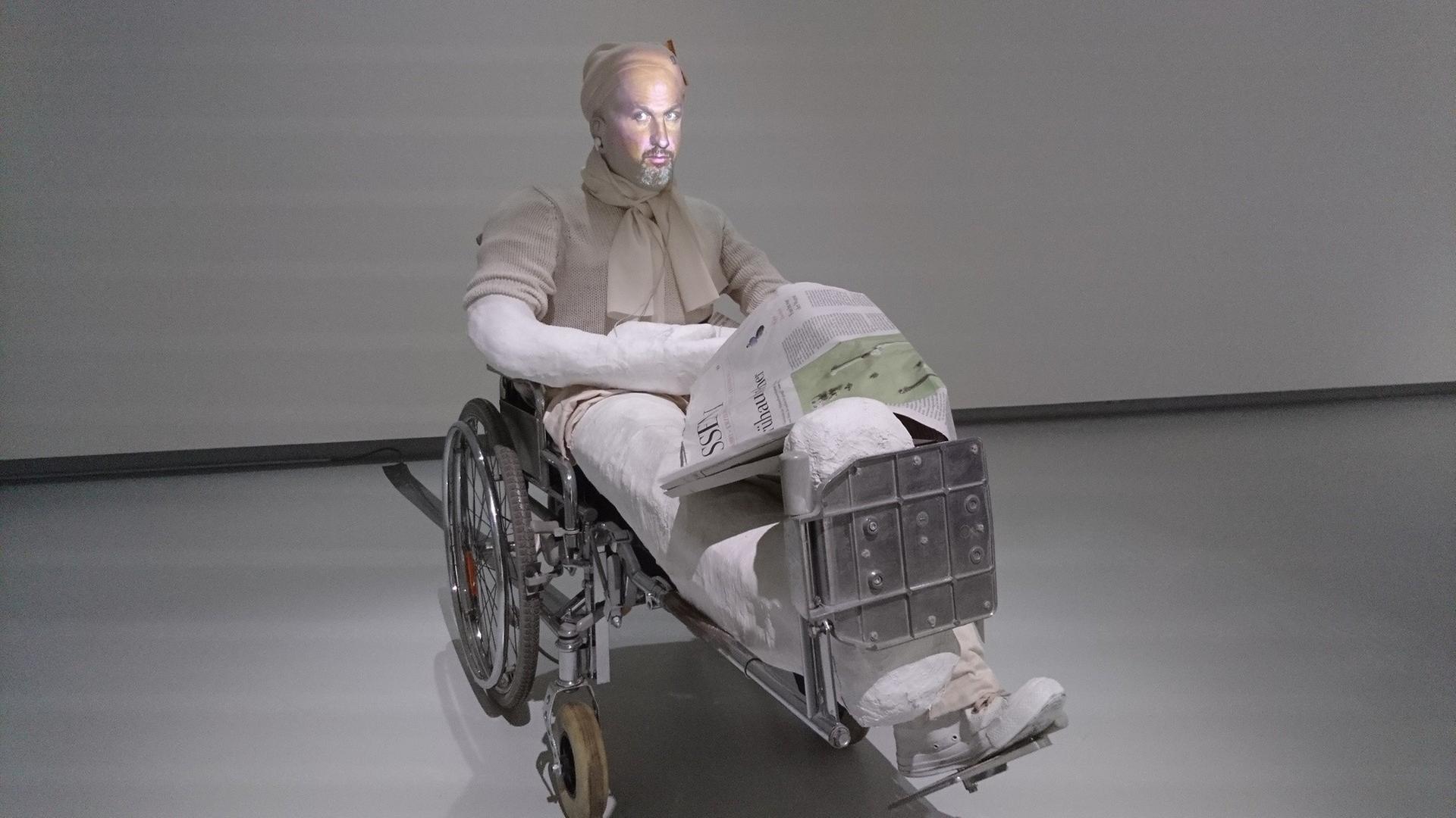 Eine in einen Rollstuhl eingegipste Puppe, auf die ein menschliches Gesicht projiziert wird. Eine in einen Rollstuhl eingegipste Puppe, auf die ein menschliches Gesicht projiziert wird.