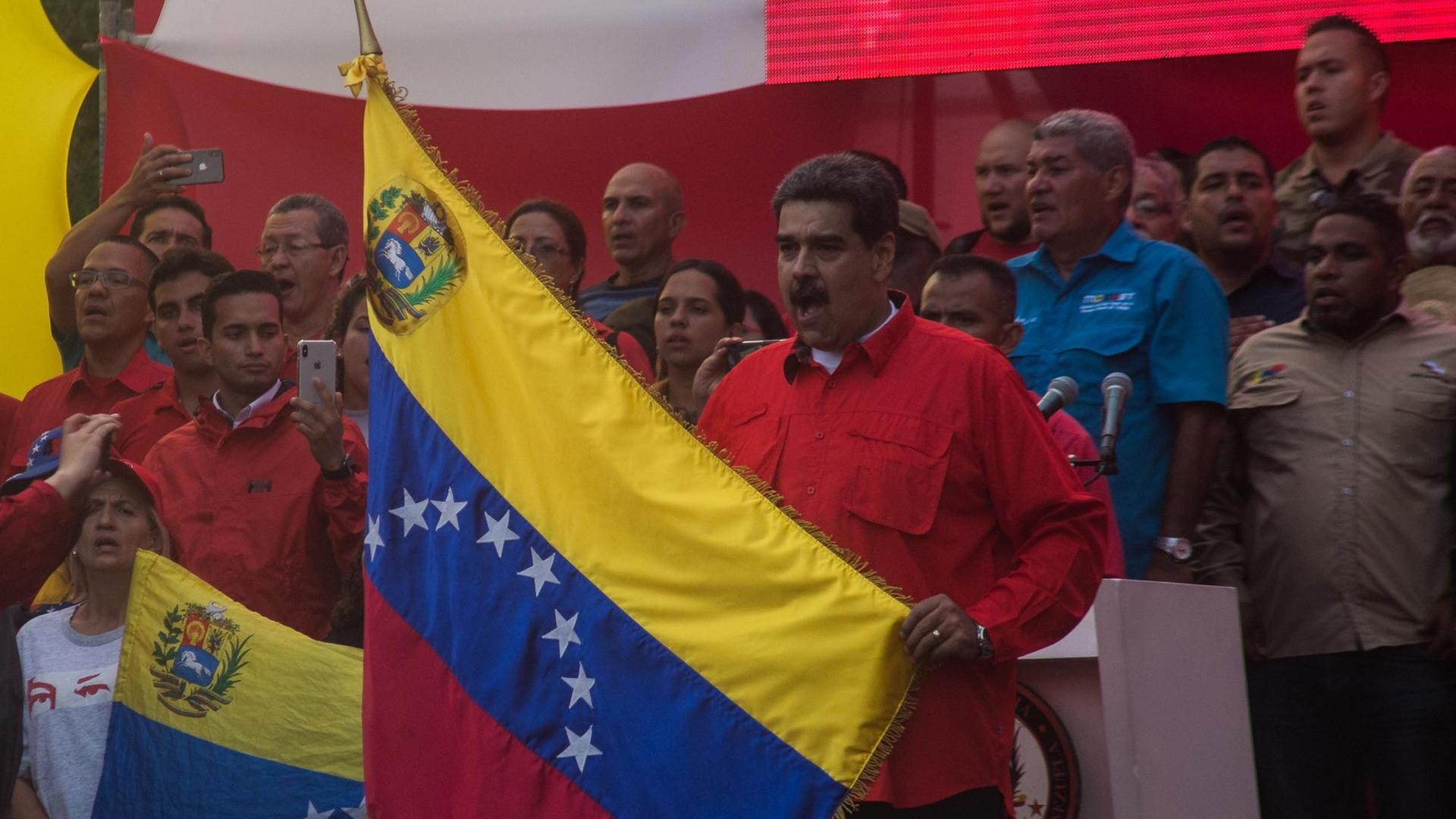 01.05.2019, Venezuela, Caracas: Nicolas Maduro (M), Präsident von Venezuela, hält während einer Kundgebung am Tag der Arbeit eine Flagge von Venezuela und singt die Nationalhymne.