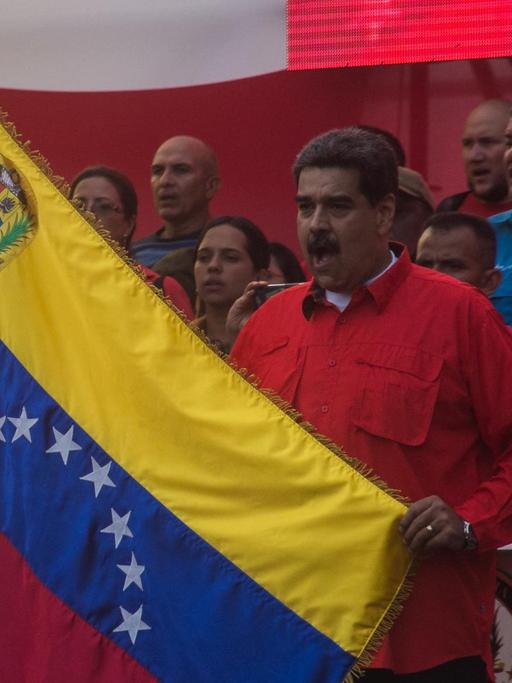 01.05.2019, Venezuela, Caracas: Nicolas Maduro (M), Präsident von Venezuela, hält während einer Kundgebung am Tag der Arbeit eine Flagge von Venezuela und singt die Nationalhymne.