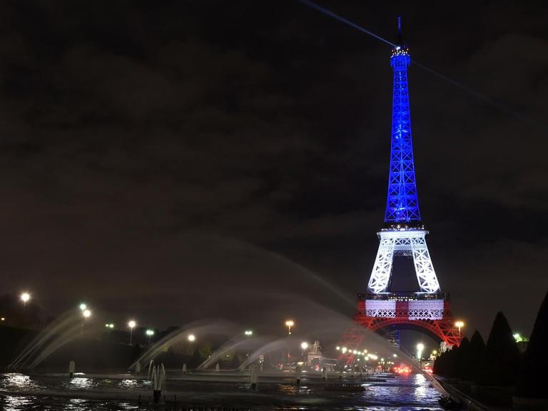 Nächtlicher Blick über die Seine auf den Eiffelturm, dessen oberer Teil blau, der mittlere Teil weiß und der untere Teil rot angestrahlt wird.