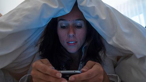 Eine Frau liegt unter der Decke im Bett und schaut auf ihr Smarphone.