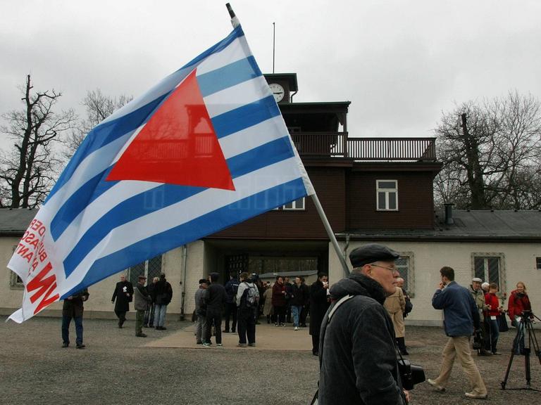 Ein Mann trägt die blau-weiß gestreifte Fahne mit rotem Wimpel durch die Gedenkstätte