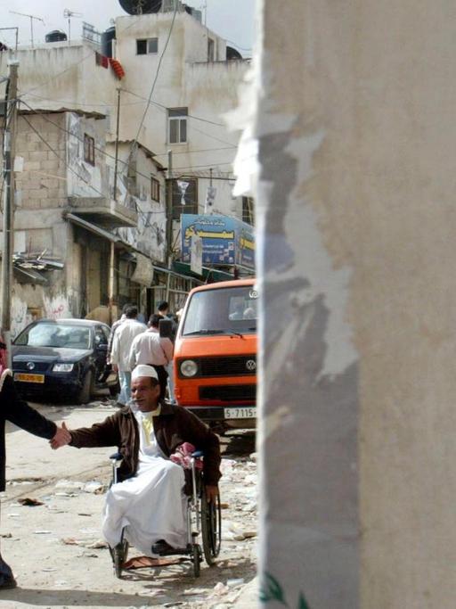 Straßenszene aus dem Flüchtlingslager al-Amari bei Ramallah. Ein Mann begrüßt einen anderen im Rollstuhl per Handschlag. Im Hintergrund eine heruntergerkommene Häuserzeile, rechts eine Hauswand mit Davidstern.