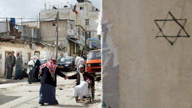 Straßenszene aus dem Flüchtlingslager al-Amari bei Ramallah. Ein Mann begrüßt einen anderen im Rollstuhl per Handschlag. Im Hintergrund eine heruntergerkommene Häuserzeile, rechts eine Hauswand mit Davidstern.