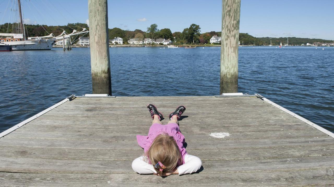 Ein Mädchen liegt entspannt an einer Mole und schaut auf ein Gewässer hinaus, auf dem Boote und Yachten sind. Ein Mädchen liegt entspannt an einer Mole und schaut auf ein Gewässer hinaus, auf dem Boote und Yachten sind.