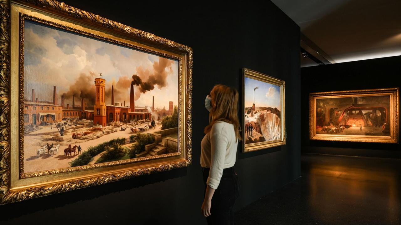 Eine Frau steht im Bucerius Kunst Forum vor Gemälden mit Industrieabbildungen. Eine Frau steht im Bucerius Kunst Forum vor Gemälden mit Industrieabbildungen.