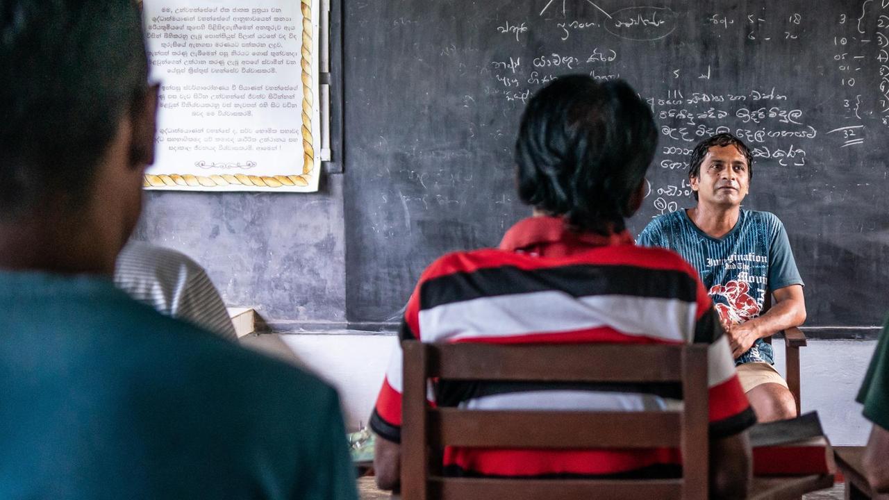 Ein Mann sitzt vor einer Tafel und spricht zu jungen Männern, die wie Schüler in einer Schule vor dem Lehrer setzen. Ein Mann sitzt vor einer Tafel und spricht zu jungen Männern, die wie Schüler in einer Schule vor dem Lehrer setzen.