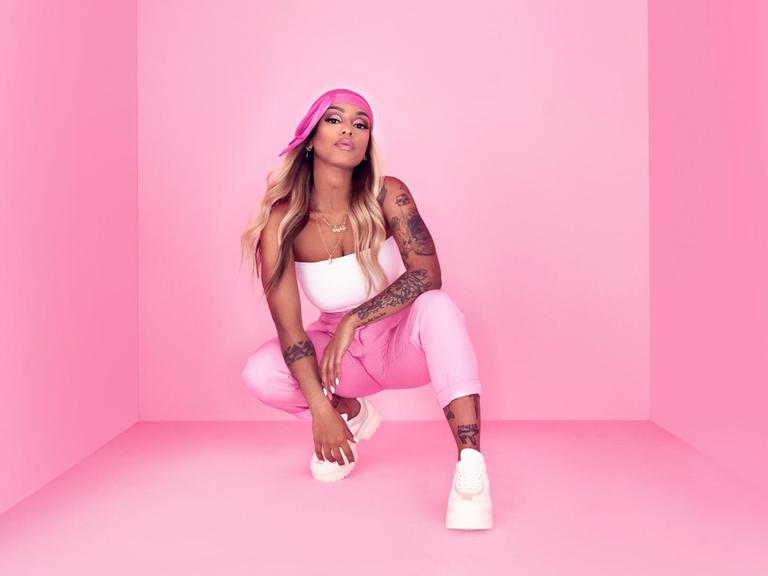 Die Rapperin Nura posiert in der Hocke vor einem pinken Hintergrund.