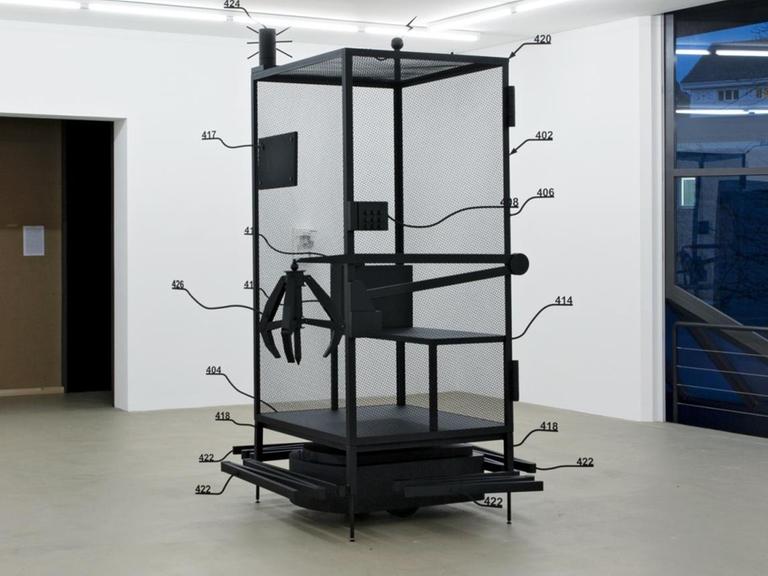 Man sieht den Prototyp eines Käfig, entwickelt für die Firma Amazon, der dann aber nicht gebaut wurde, im Ausstellungsraum, in dem die Arbeiter sitzen sollten. Der Käfig hat einen Greifarm an der Seite und Rollen drunter.