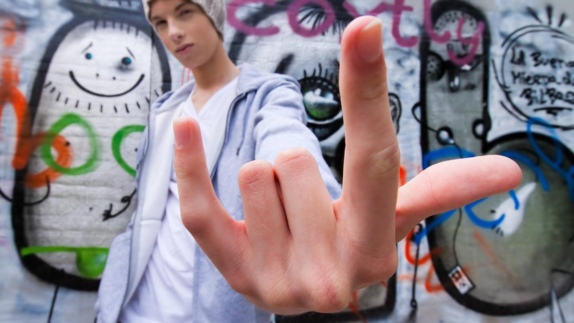 Ein cool blickender jugendlicher Mann gestikuliert vor einer Wand mit Graffiti Ein cool blickender jugendlicher Mann gestikuliert vor einer Wand mit Graffiti