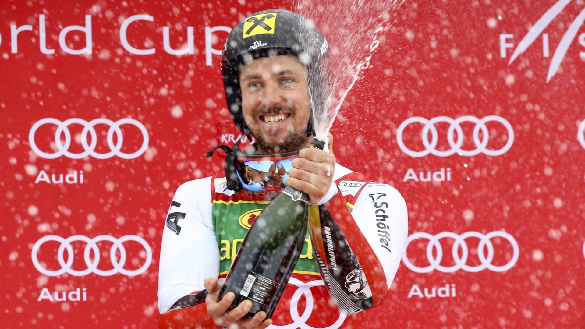 Hirscher steht lachend mit Helm vor einer roten Wand mit den Sponsorenlogos und schüttelt eine Sektflasche. Hirscher steht lachend mit Helm vor einer roten Wand mit den Sponsorenlogos und schüttelt eine Sektflasche.