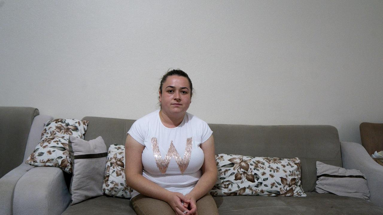 Eine junge Frau mit Pferdeschwanz und in weißem T-Shirt sitzt in einer Wohnung auf einem beigen Sofa. Eine junge Frau mit Pferdeschwanz und in weißem T-Shirt sitzt in einer Wohnung auf einem beigen Sofa.
