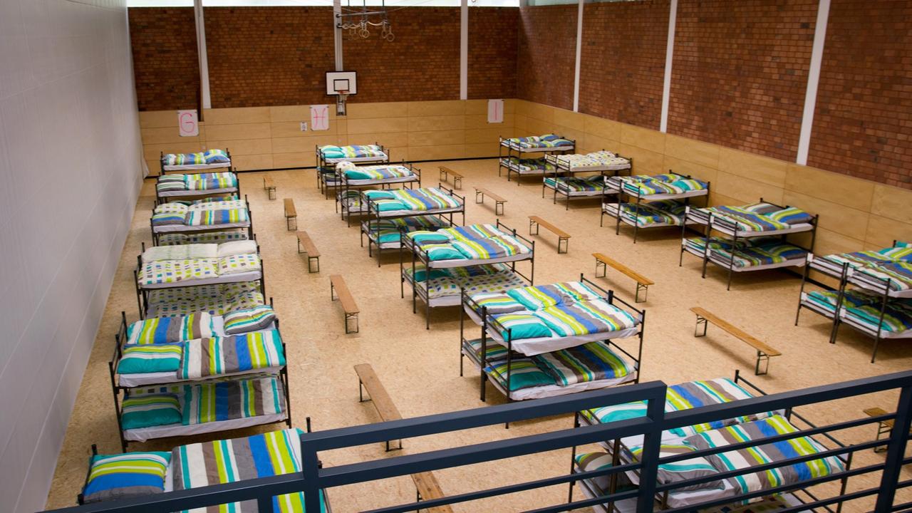 Eine umfunktionierte Turnhalle, in der in drei Reihen Betten stehen. Eine umfunktionierte Turnhalle, in der in drei Reihen Betten stehen.