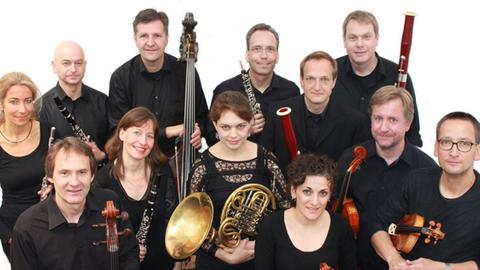 Zwölf Menschen in schwarzer Konzertkleidung stehen eng zusammen und blicken leicht nach oben in die Kamera, dabei hält jeder sein/ihr Instrument in der Hand vor sich.