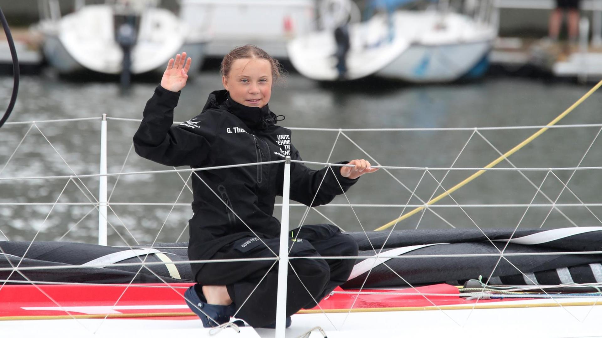 Greta Thunberg kniet an Bord, hält sich an der Reling fest und winkt lächelnd in die Kamera. Greta Thunberg kniet an Bord, hält sich an der Reling fest und winkt lächelnd in die Kamera.