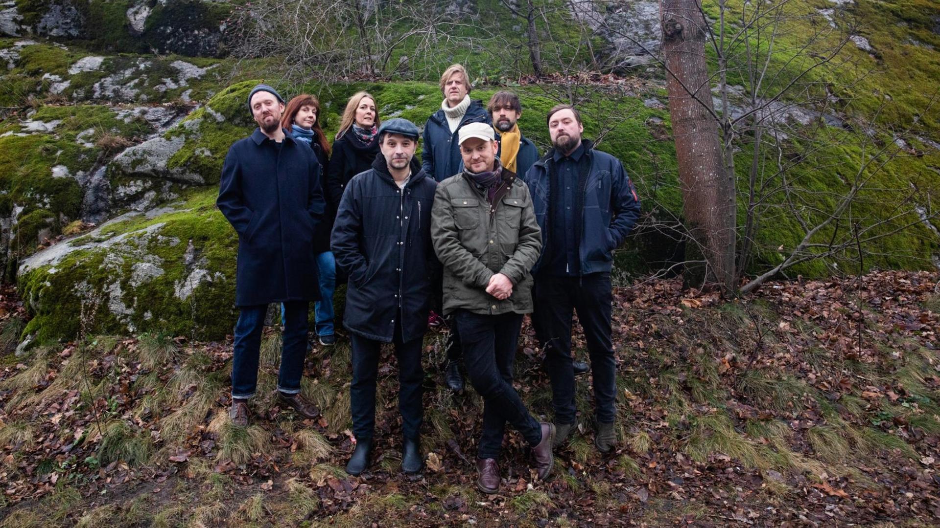 Eine Gruppe von acht Menschen (zqwi Frauen, sechs Männer) steht in freier Natur vor moosbewachsenen Felden, blätterlose Bäume im Hintergrund. Eine Gruppe von acht Menschen (zqwi Frauen, sechs Männer) steht in freier Natur vor moosbewachsenen Felden, blätterlose Bäume im Hintergrund.