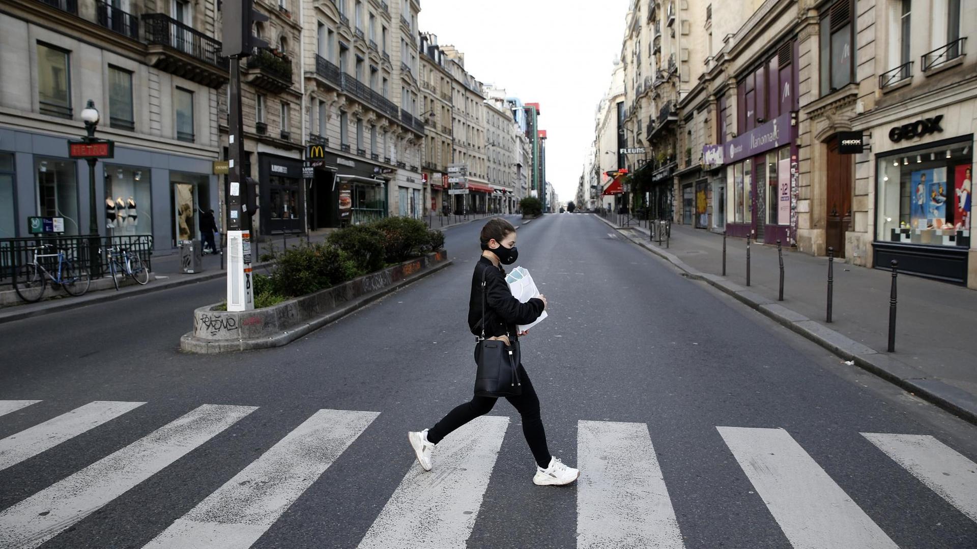 Eine junge Frau mit Atemschutzmaske überquert eine ansonsten völlig leere Straße in Paris. Sämtliche Geschäfte sind geschlossen. Eine junge Frau mit Atemschutzmaske überquert eine ansonsten völlig leere Straße in Paris. Sämtliche Geschäfte sind geschlossen.