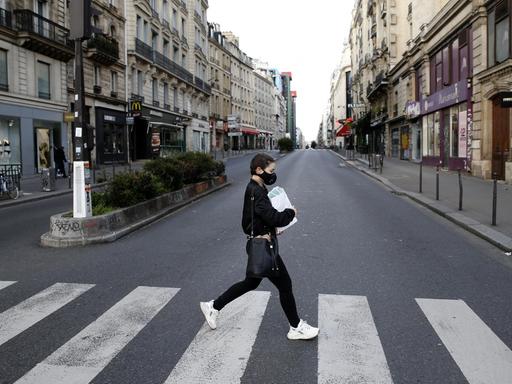 Eine junge Frau mit Atemschutzmaske überquert eine ansonsten völlig leere Straße in Paris. Sämtliche Geschäfte sind geschlossen.