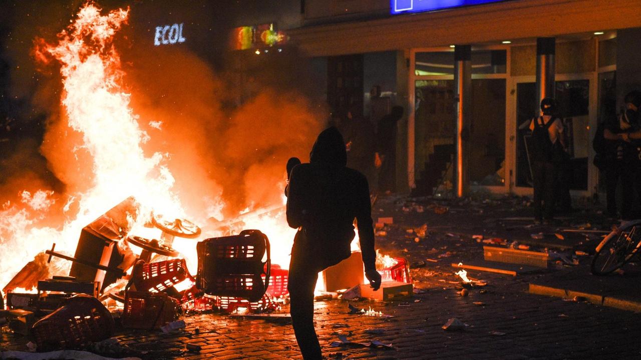Vor dem Supermarkt mit zerborstenen Scheiben brennt im Dunkeln die Barrikade; ein Demonstrant kickt etwas in das Feuer.