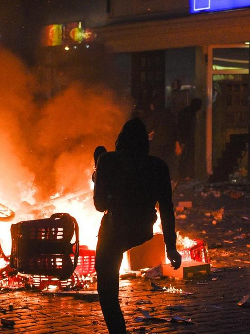 Vor dem Supermarkt mit zerborstenen Scheiben brennt im Dunkeln eine Barrikade; ein Demonstrant kickt etwas in das Feuer.