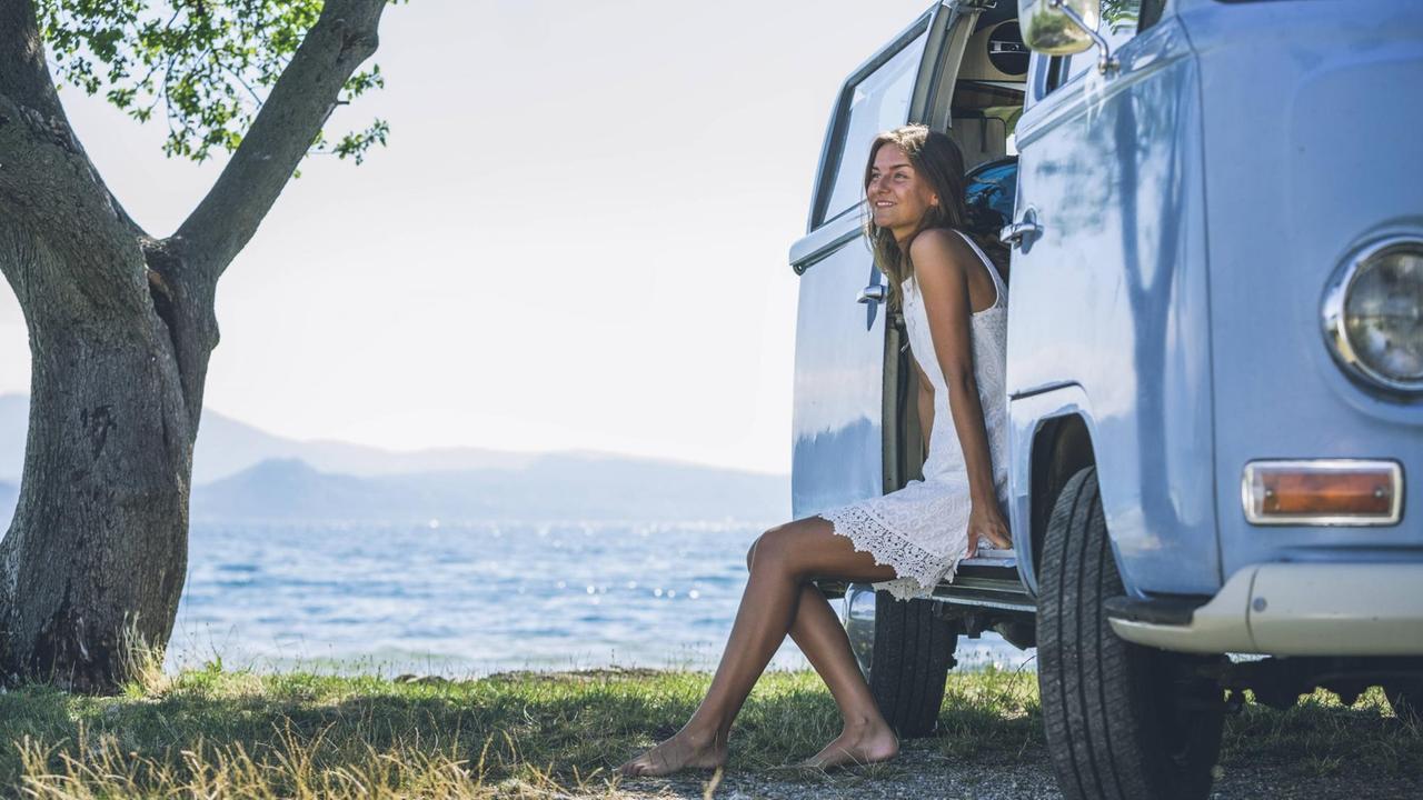 Eine Frau sitzt an ihrem Campingbus am Wasser und genießt die Sonne. Eine Frau sitzt an ihrem Campingbus am Wasser und genießt die Sonne.