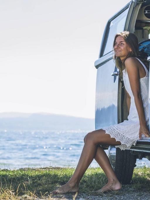 Eine Frau sitzt an ihrem Campingbus am Wasser und genießt die Sonne.