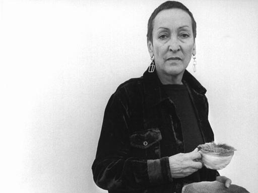 Die Künstlerin Meret Oppenheim 1975 mit eigenen Werken: Tasse und Teller mit Pelz überzogen und ein Kohlenobjekt