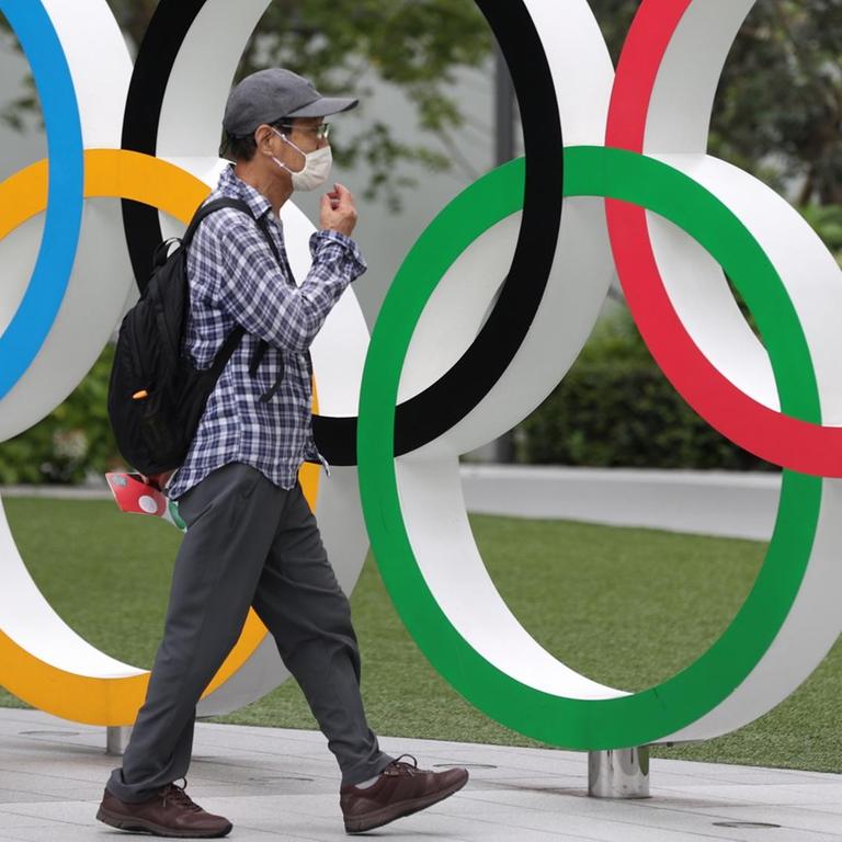 Ein Mann mit Maske läuft an einer Installation Olympischer Ringe vorbei, die in Tokio an die wegen Corona verschobene Sportveranstaltung erinnert.  