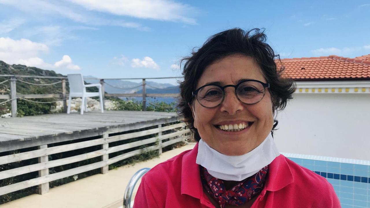 Eine dunkelhaarige Frau mit Brille und einem weißen Mundschutz unterm Kinn lächelt im pinkroten Hemd in die Kamera. Im Hintergrund sind Meer und Berge zu erkennen. Eine dunkelhaarige Frau mit Brille und einem weißen Mundschutz unterm Kinn lächelt im pinkroten Hemd in die Kamera. Im Hintergrund sind Meer und Berge zu erkennen.