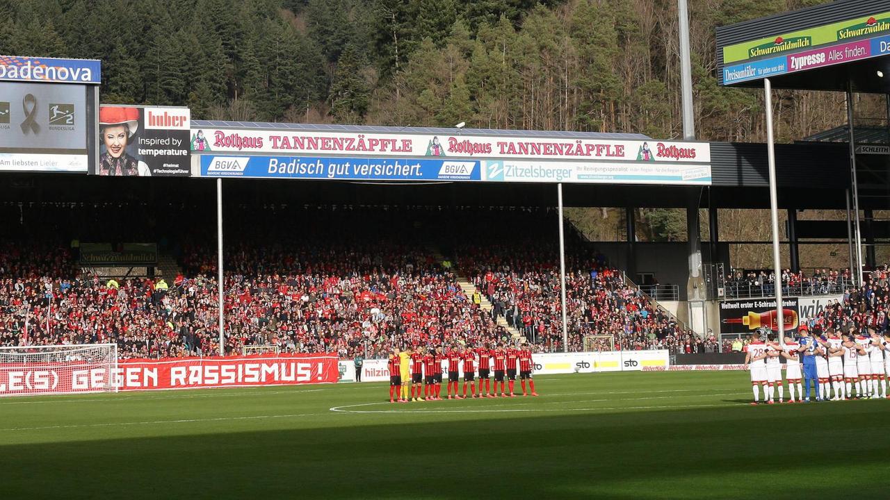 Zu sehen ist das Freiburger-Fußball-Stadion. Auf dem Rasen stehen in einem Halbkreis die Spieler von Freiburg und Düsseldorf, auf der Leinwand wird den Opfern des Anschlags in Hanau gedacht. Zu sehen ist das Freiburger-Fußball-Stadion. Auf dem Rasen stehen in einem Halbkreis die Spieler von Freiburg und Düsseldorf, auf der Leinwand wird den Opfern des Anschlags in Hanau gedacht.