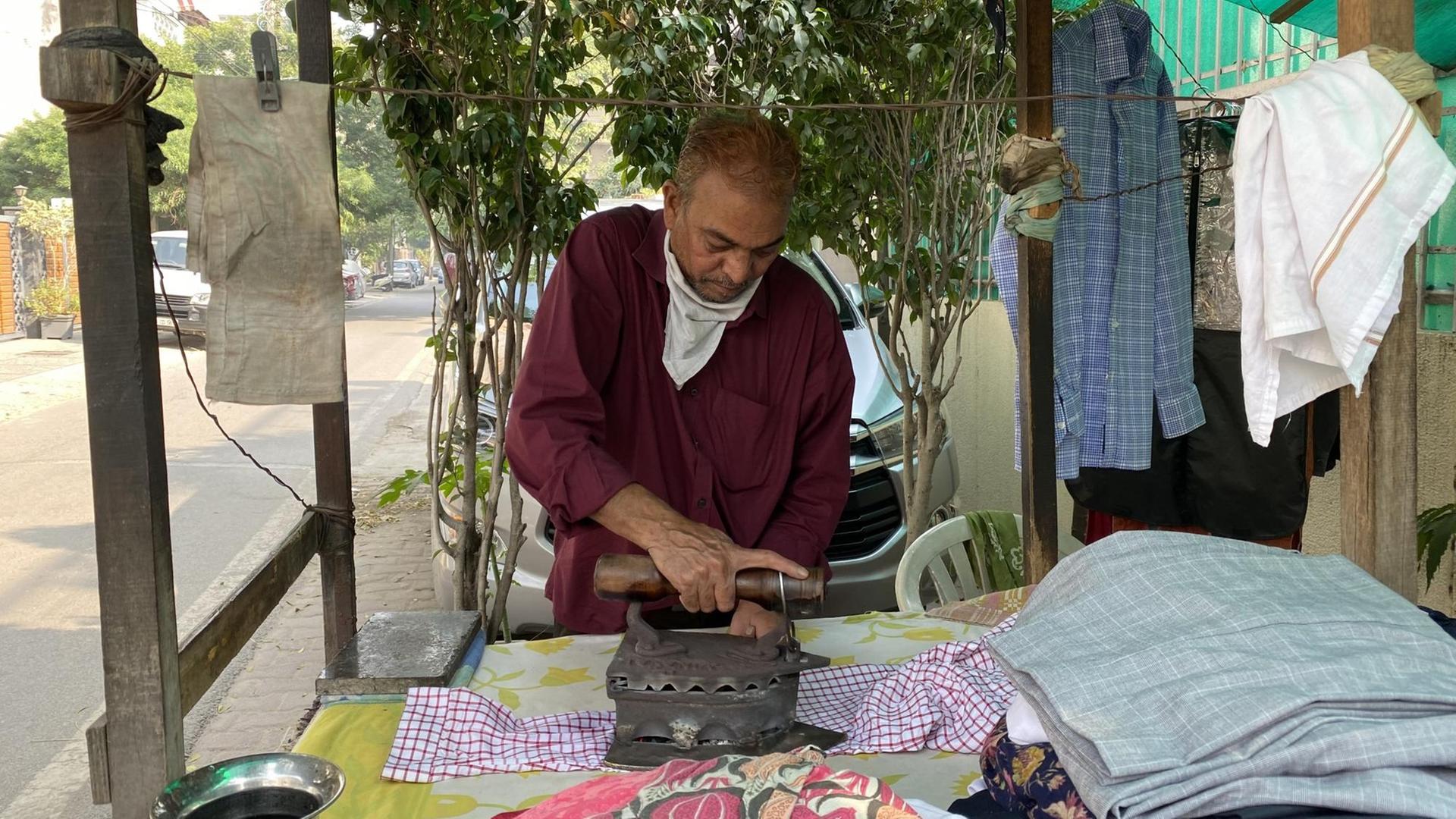 Ein Mann im roten Hemd bügelt mit einem schweren altmodischen Bügeleisen an einer Straße Kleidung.