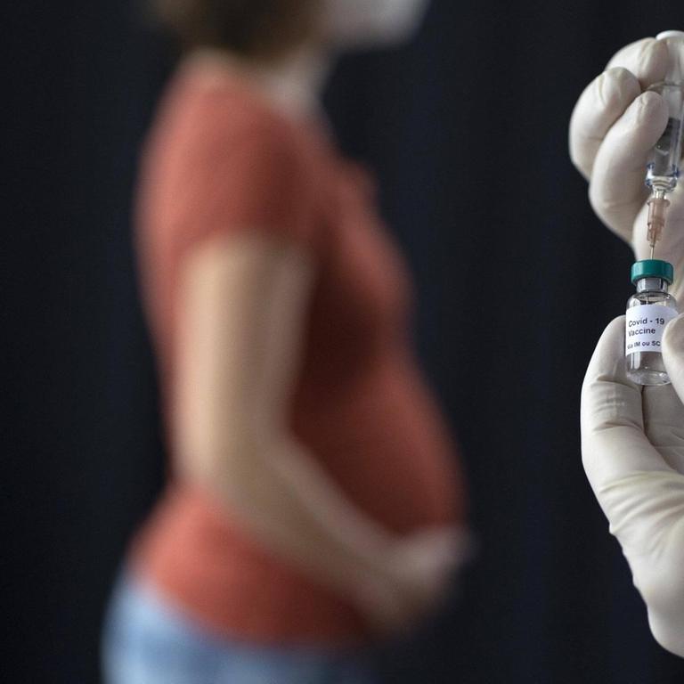 Eine Schwangere hält sich den Bauch; im Vordergrund sind zwei Hände zu sehen, die Impfstoff aus einer Dose in eine Spritze ziehen. (Symboldbild)